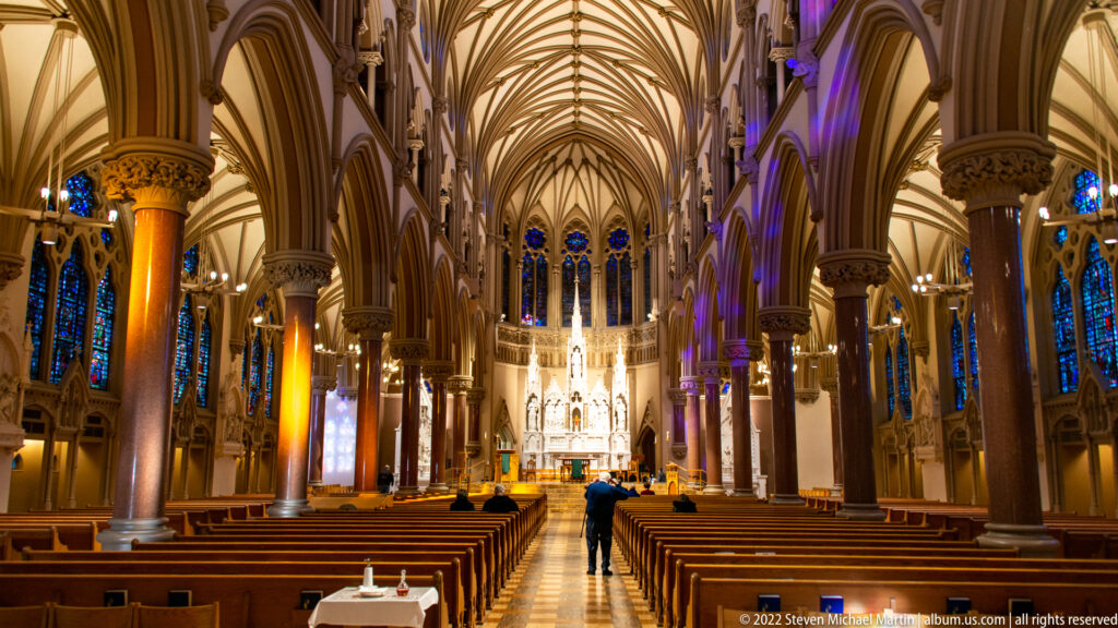Central aisle leads through a vaulted cathedral nave toward a bright altar, with pews and stained glass.