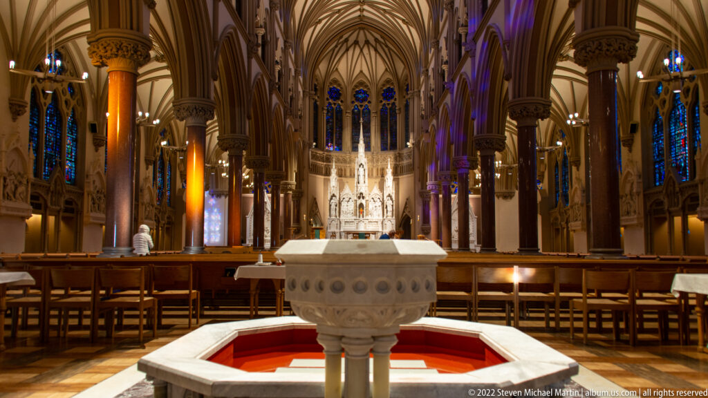 Stone baptismal font stands before rows of pews and the distant cathedral altar.