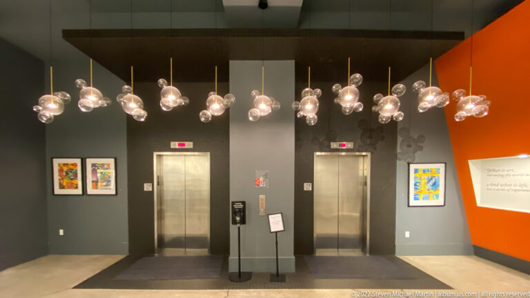 Glass globe pendants hang above two elevators, framed art, and a sign in a modern lobby.