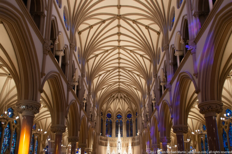Upward view of pale rib vaults and pointed arches above columns and blue stained-glass windows.