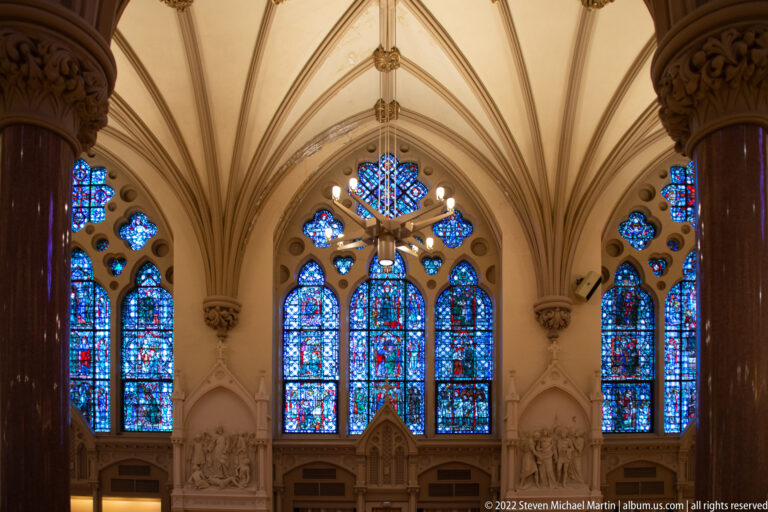 Crucifix hangs before a central stained-glass window between narrower lancet windows above the sanctuary.