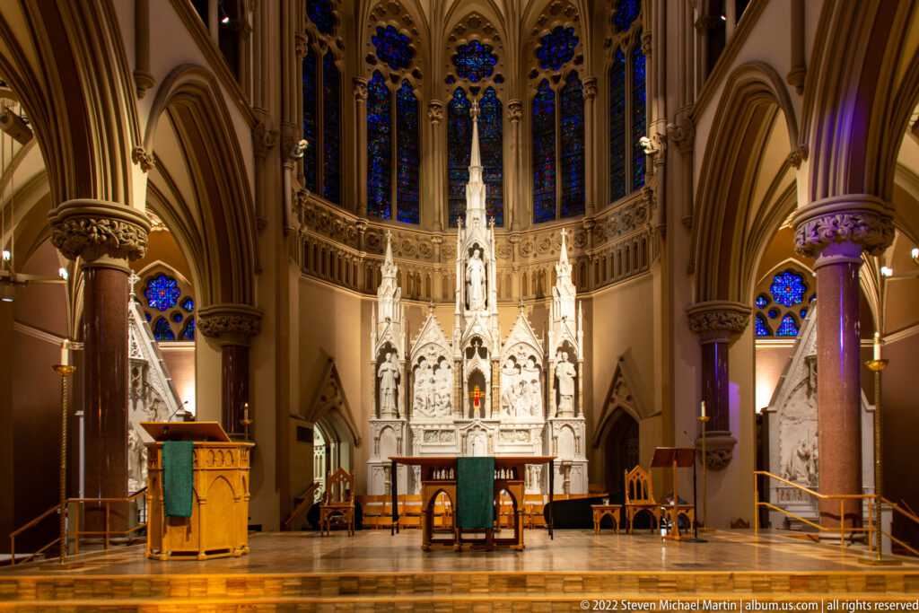 White sculpted altar stands beneath blue stained glass, with a lectern and chairs across the sanctuary.