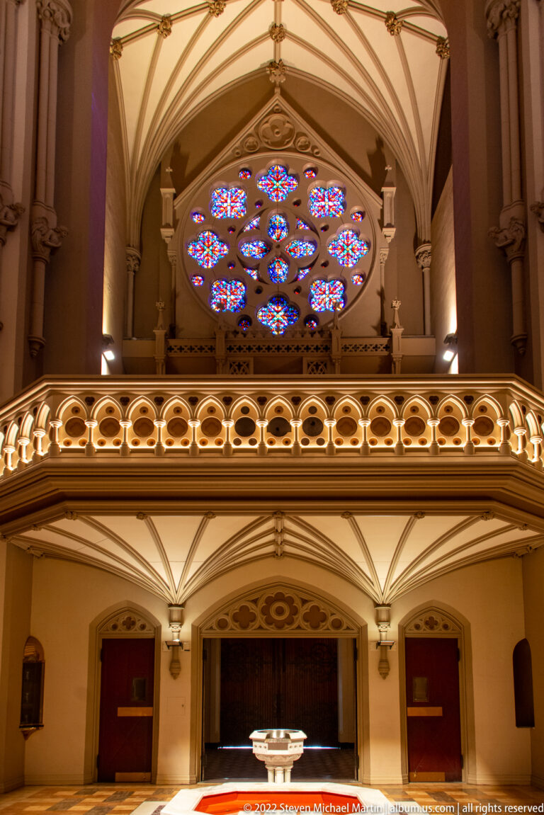 Upper gallery and patterned balustrade beneath a glowing rose window, with a lower room visible below.