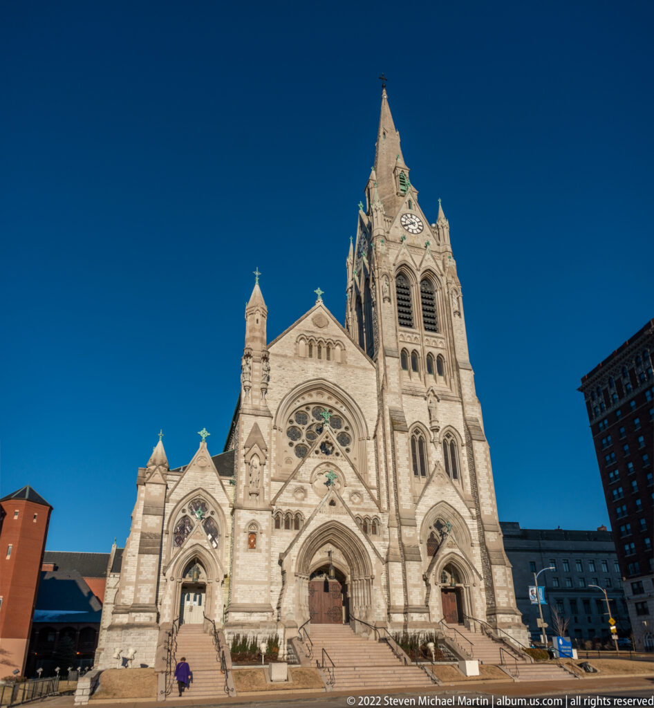 Pale stone cathedral façade with central tower, three portals, circular window and broad steps.