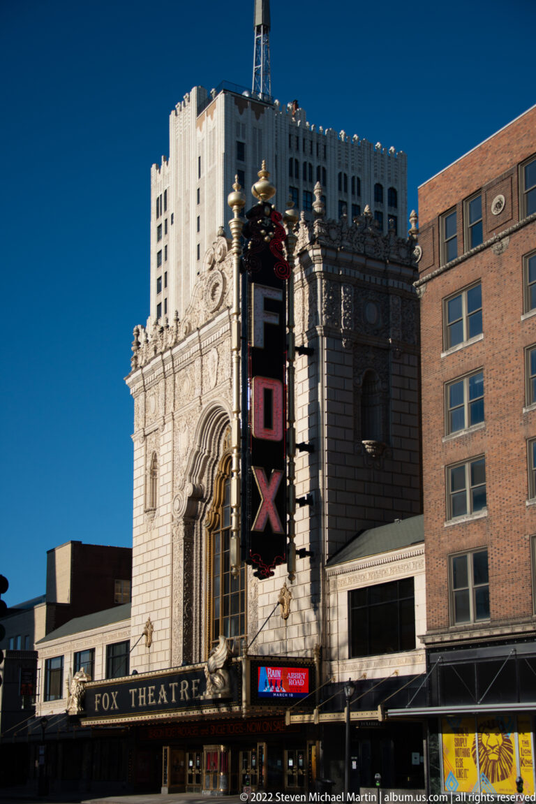 Fox Theatre and vertical sign framed by adjoining downtown buildings beneath a blue sky.