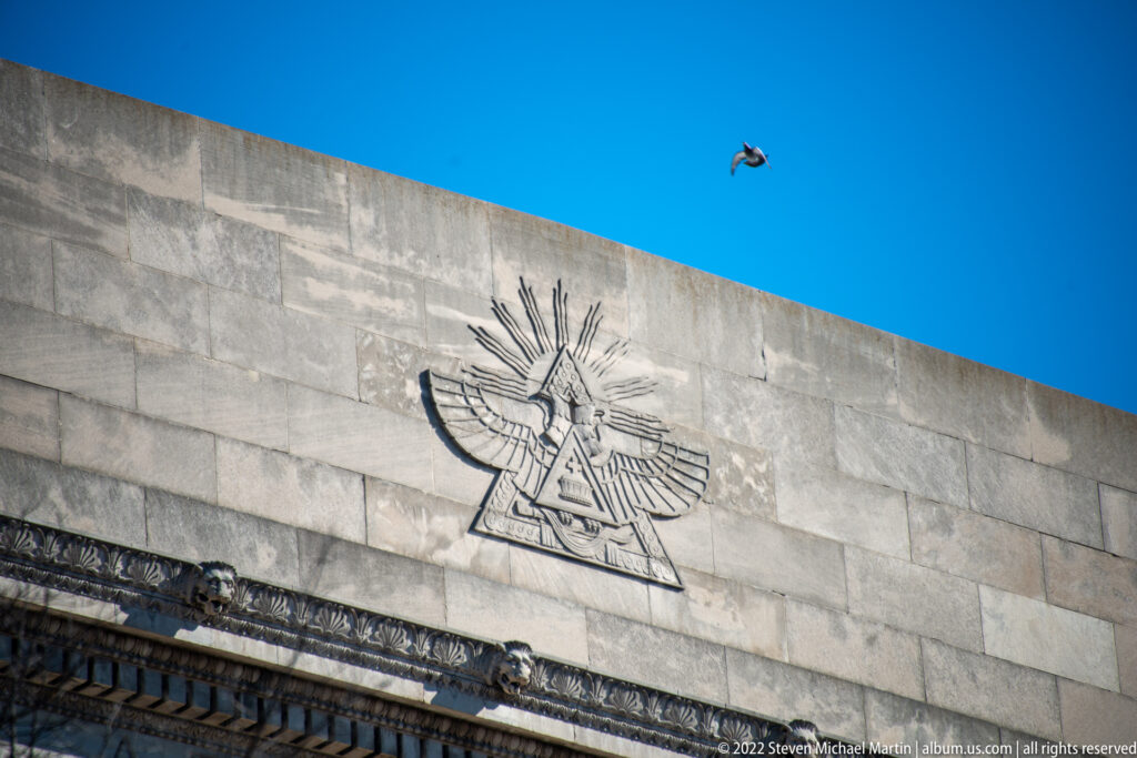 Stone wall relief of a winged figure beneath a small bird crossing the blue sky.