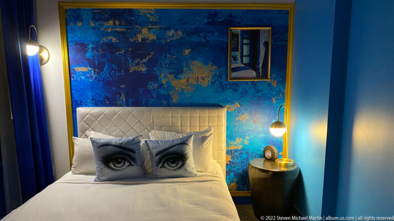 A white bed with eye-patterned pillows stands before a blue mural wall, framed artwork, and a bedside lamp.