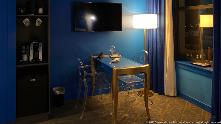 A blue desk and chair sit beside a curtained window beneath a television in a dim room.
