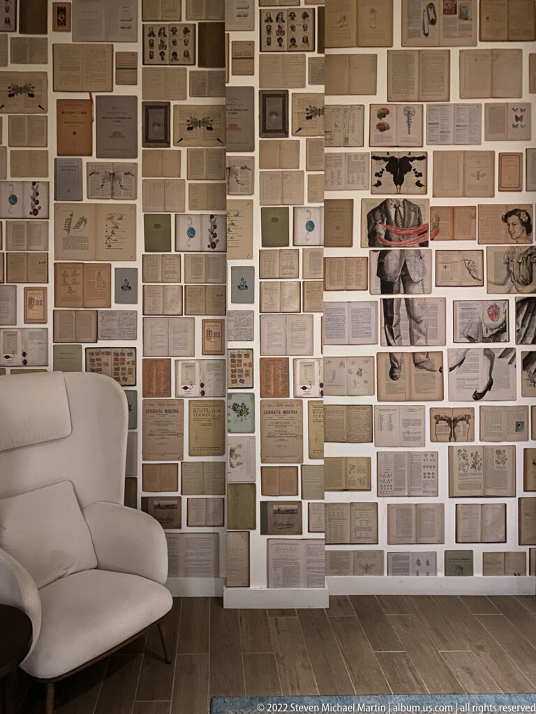 A white lounge chair faces a wall densely covered with paper fragments, photographs, text, and diagrams.
