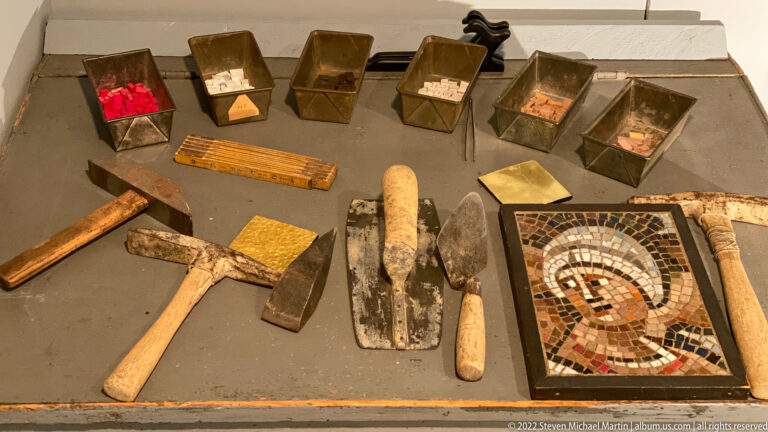 Mosaic tools and material samples are displayed beside a small framed mosaic portrait.