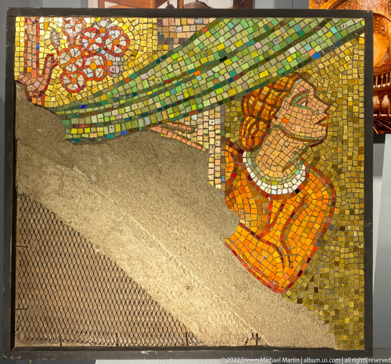 A partly completed mosaic shows a figure in orange and red amid green tesserae and exposed backing.