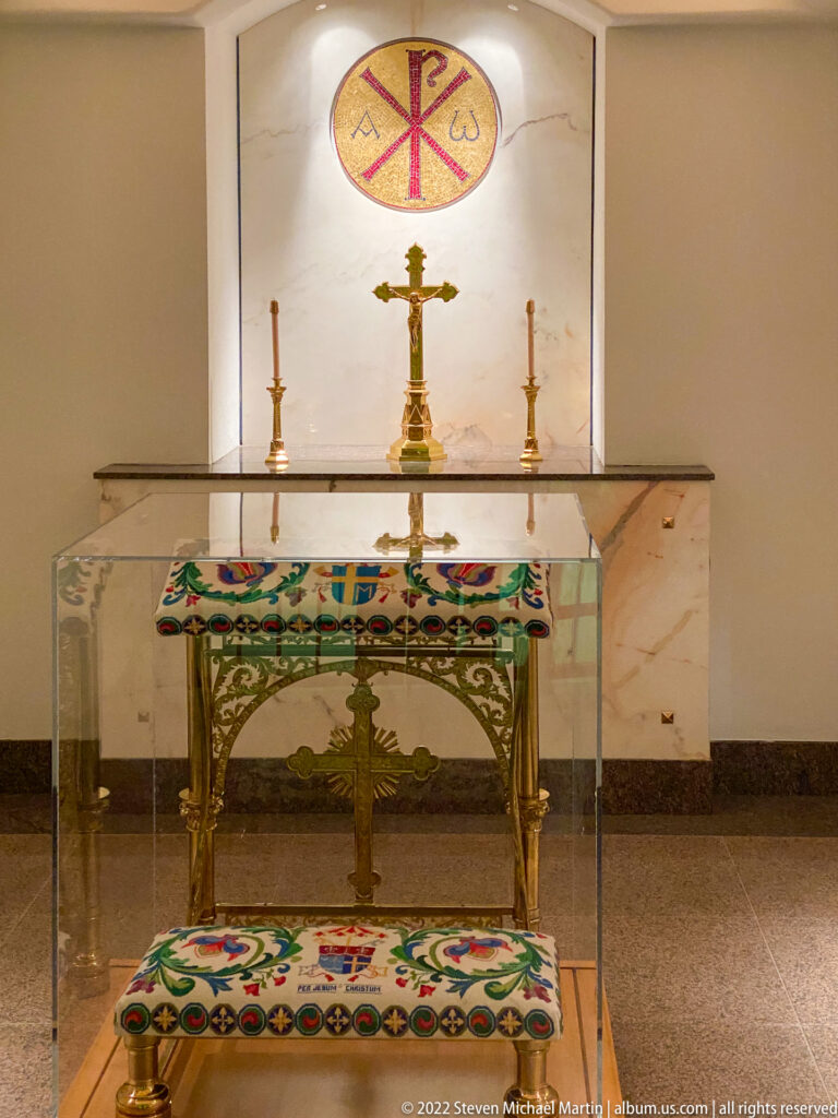 An embroidered altar cloth sits inside a clear case beneath a wall-mounted Chi-Rho emblem and crucifix.