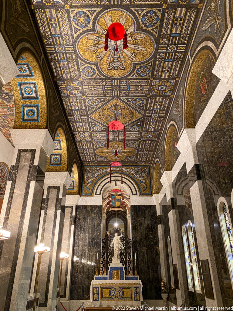 A cathedral nave extends toward the altar beneath a patterned mosaic ceiling, with marble piers and rows of pews.