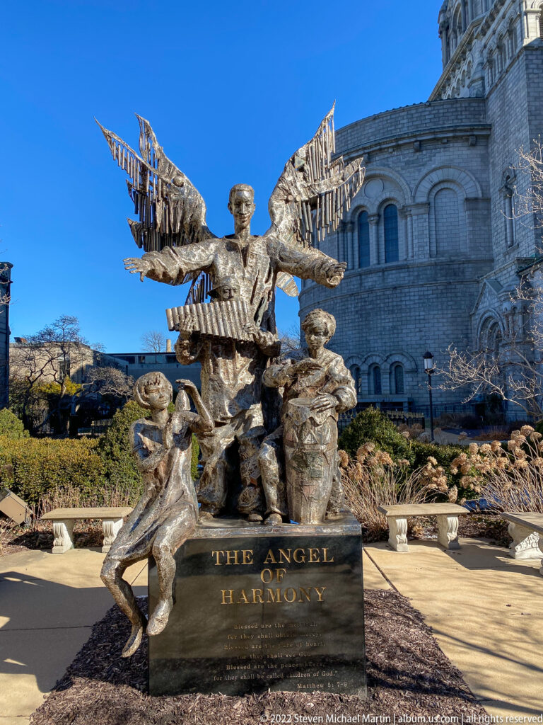 A bronze winged figure rises above two smaller figures on a pedestal labeled “The Angel of Harmony,” beside the cathedral façade.