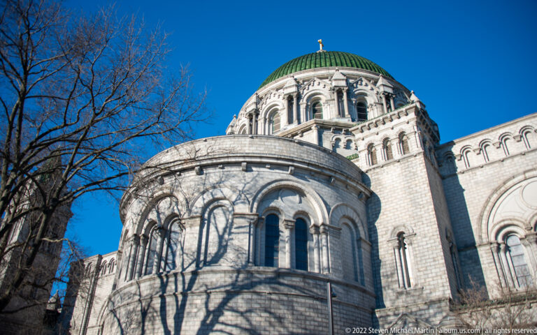 The cathedral’s rounded stone apse and arched windows rise beneath a green dome, with tree shadows crossing the façade.