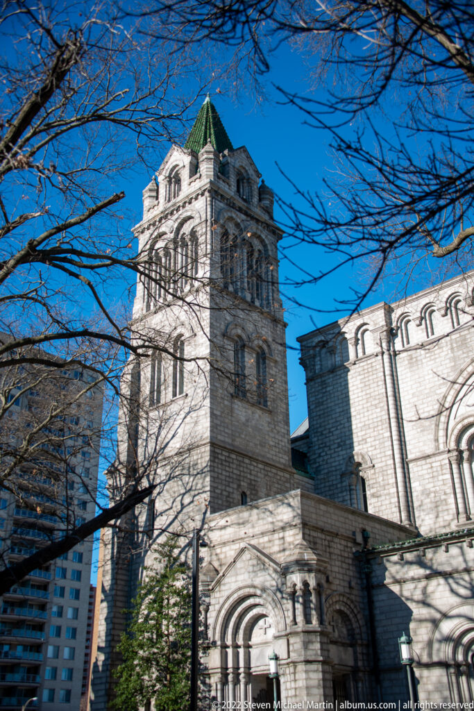A green-roofed stone bell tower rises above the cathedral walls behind bare branches.