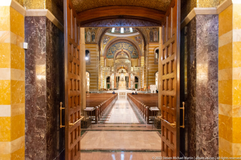 Open wooden doors frame the cathedral’s central aisle, leading through the nave toward the illuminated altar.