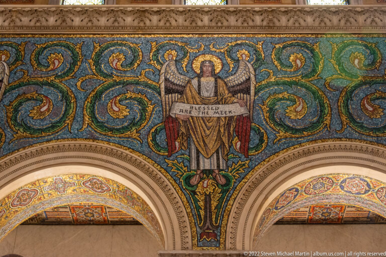 A mosaic angel holds a scroll reading “Blessed are the meek” above two arches amid green scrolling foliage.
