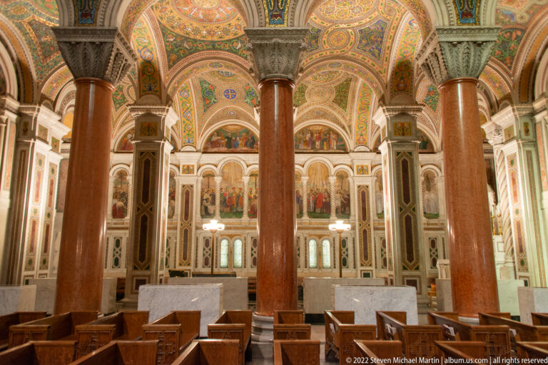 Rows of pews sit beneath mosaic arches and stained-glass windows between tall reddish marble columns.