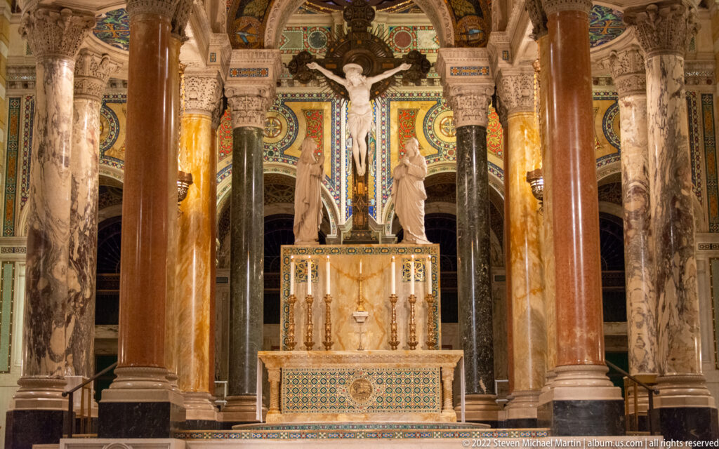A crucifix stands above the altar between marble columns and statues against a mosaic-decorated sanctuary wall.