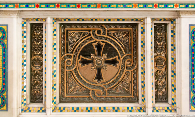 A bronze door panel with a central cross and scrolling ornament is framed by blue, yellow, and green tiles.