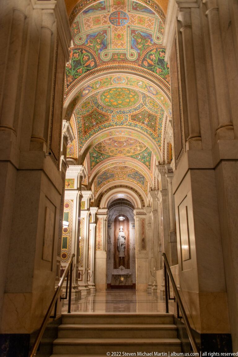 Stone steps and handrails lead through a mosaic-ceiling passage toward a statue in a distant niche.