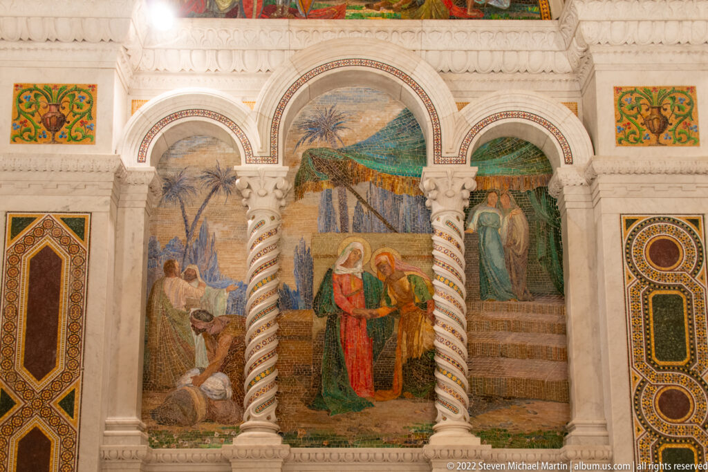 A biblical narrative mosaic fills three arches in a white marble wall, with robed figures, palm trees, patterned columns, and geometric borders.