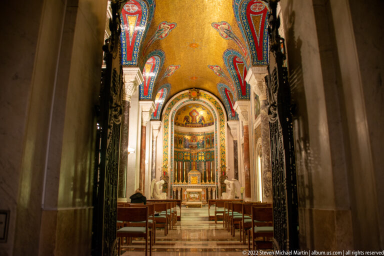 A dark doorway frames a chapel aisle lined with pews, leading toward a brightly lit mosaic altar beneath a patterned ceiling.