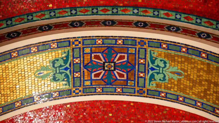 A blue, red, and green geometric medallion sits in a gold mosaic field bordered by curved bands of patterned tesserae.