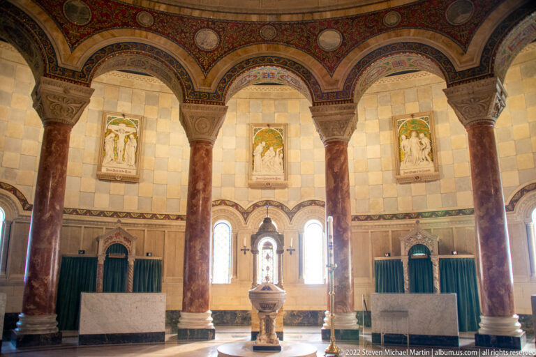 A stone baptismal font stands before three arched bays, between marble columns and beneath framed religious reliefs.