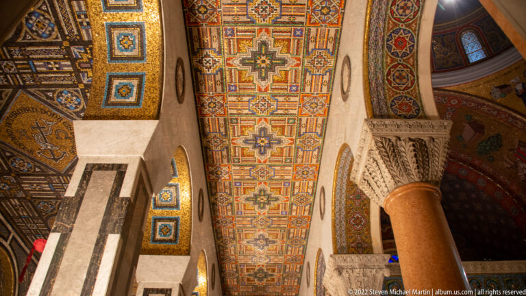 An upward view follows a long multicolored mosaic ceiling bay between stone piers, carved capitals, and patterned arches.