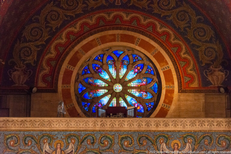 A circular blue and red rose window rises above a carved choir gallery, surrounded by a painted arch and mosaic band.