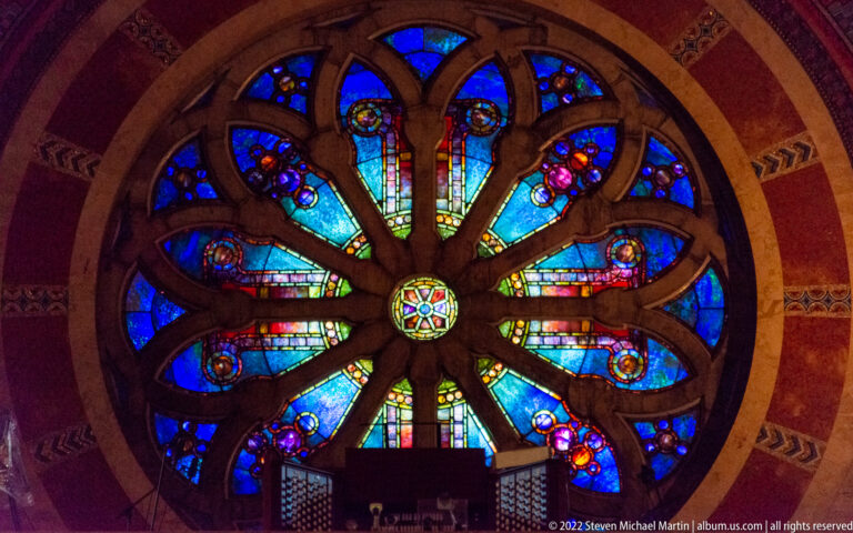 A close view of a rose window shows blue glass petals, turquoise rays, colored roundels, and dark radial tracery.
