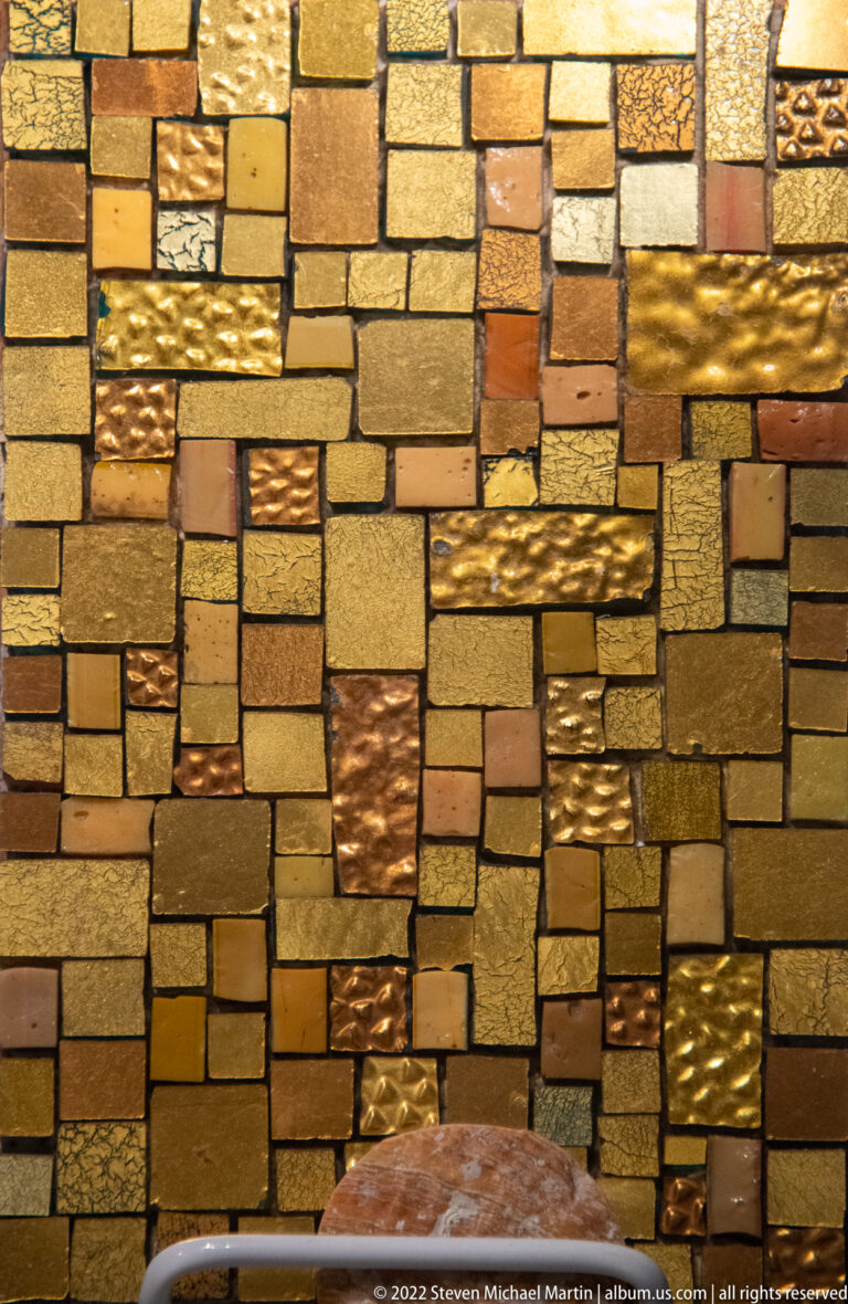Small gold, amber, and bronze tesserae cover a dense wall section, with varied textures and a curved dark edge below.