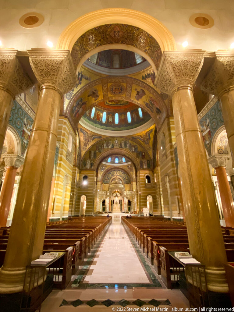 A low centered view looks down the aisle between pews toward the altar and mosaic-covered apse.