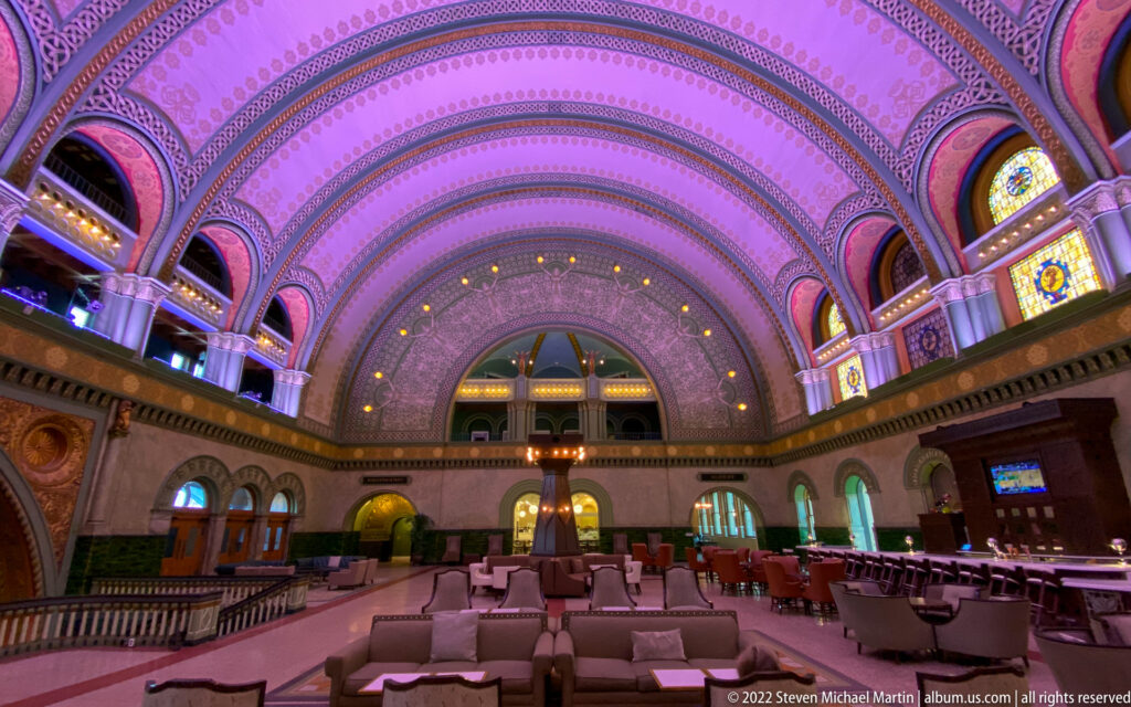 Purple light washes the vaulted Grand Hall above lounge seating, balconies, and an arched window.