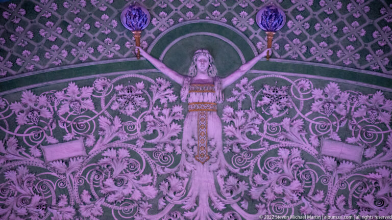 A sculpted female figure raises her arms amid an ornate wall of leaves and curling forms.