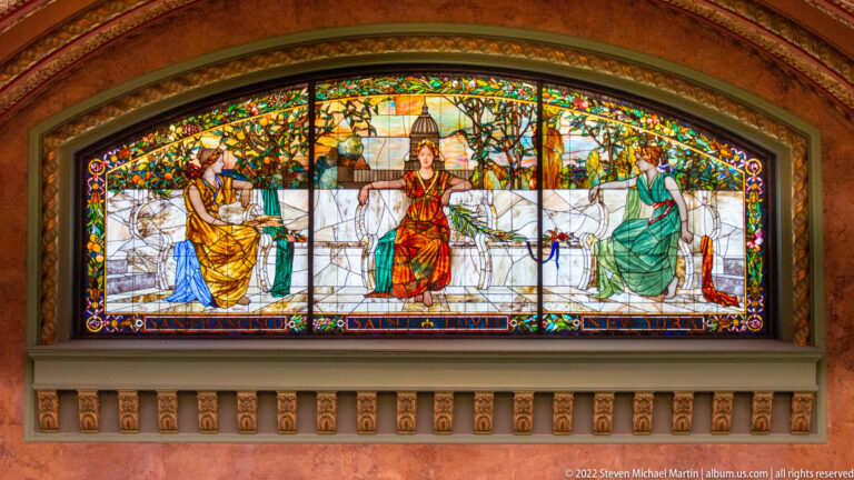 Three seated figures appear in a broad stained-glass window with a colorful border.