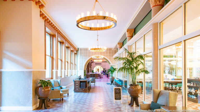 A bright corridor with circular chandeliers leads toward an arch past windows, seating, and plants.