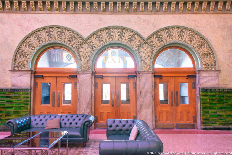 Three wooden doors beneath carved arches stand behind dark sofas; “EXIT” appears over the center door.