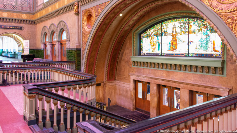 Several balconies overlook doors and a large stained-glass panel with seated figures beyond.