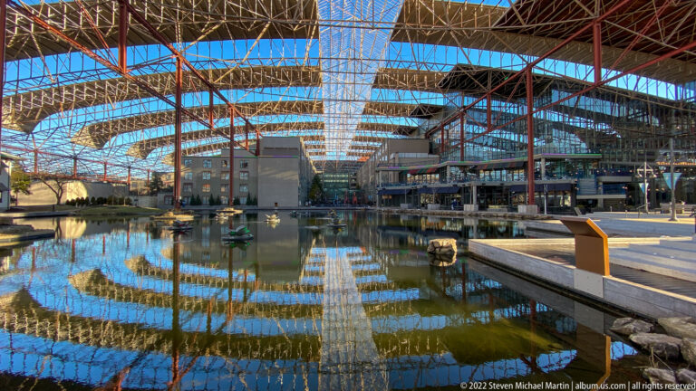 Iron roof trusses span a reflecting pool, walkways, and planters inside the former train shed.