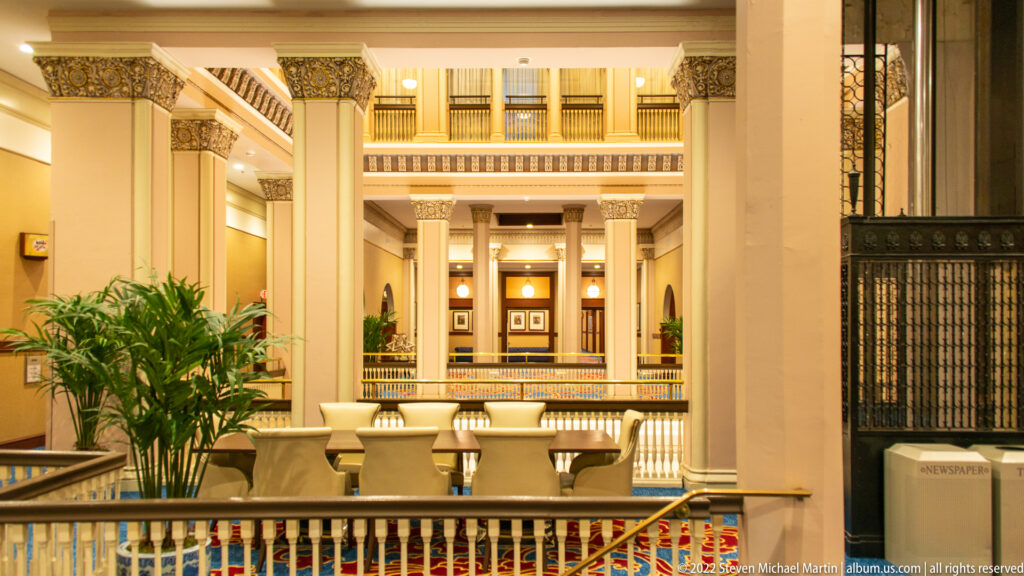 Interior balconies overlook tables and chairs, with a plant and seating area in the foreground.