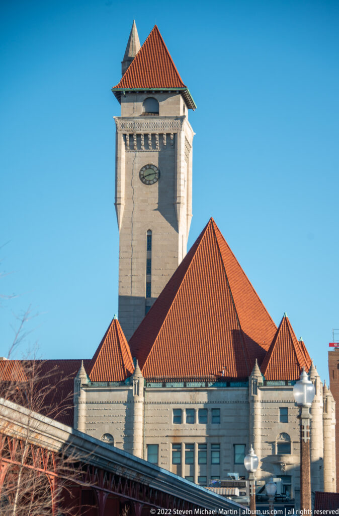 A stone clock tower rises behind steep red-tiled roofs, with a steel truss crossing the foreground.