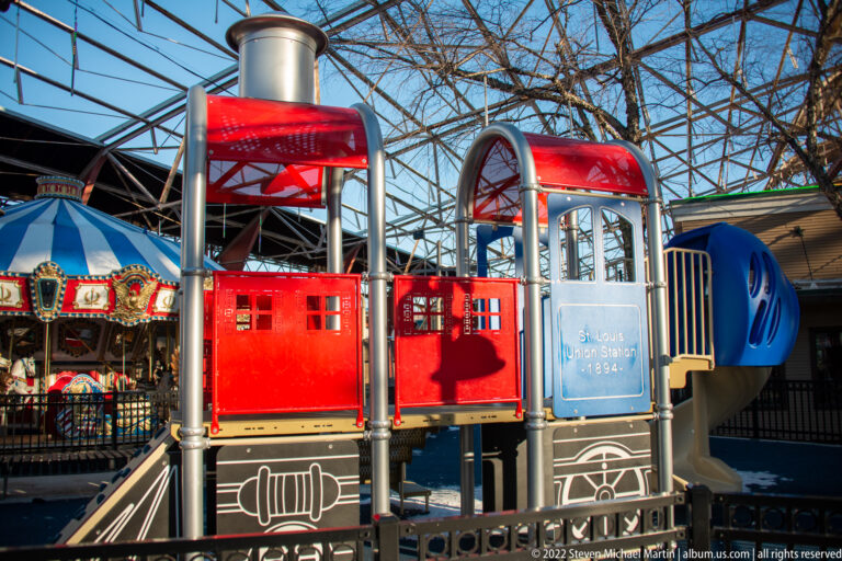 Red and blue ticket booths stand in front of a decorated carousel beneath steel roof trusses.