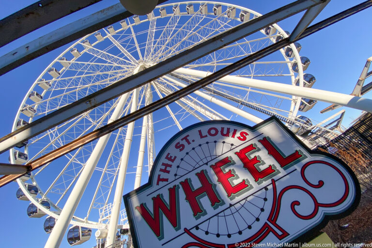 A sign reading “The St. Louis Wheel” stands beneath the large white Ferris wheel.