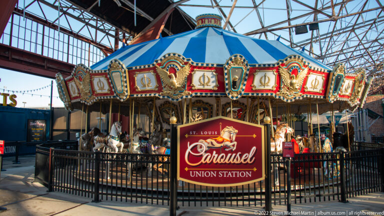 A colorful carousel with carved horses and a blue canopy stands behind a black fence indoors.