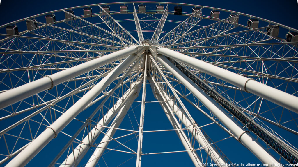 White support legs and intersecting spokes converge at the center of a Ferris wheel against blue sky.