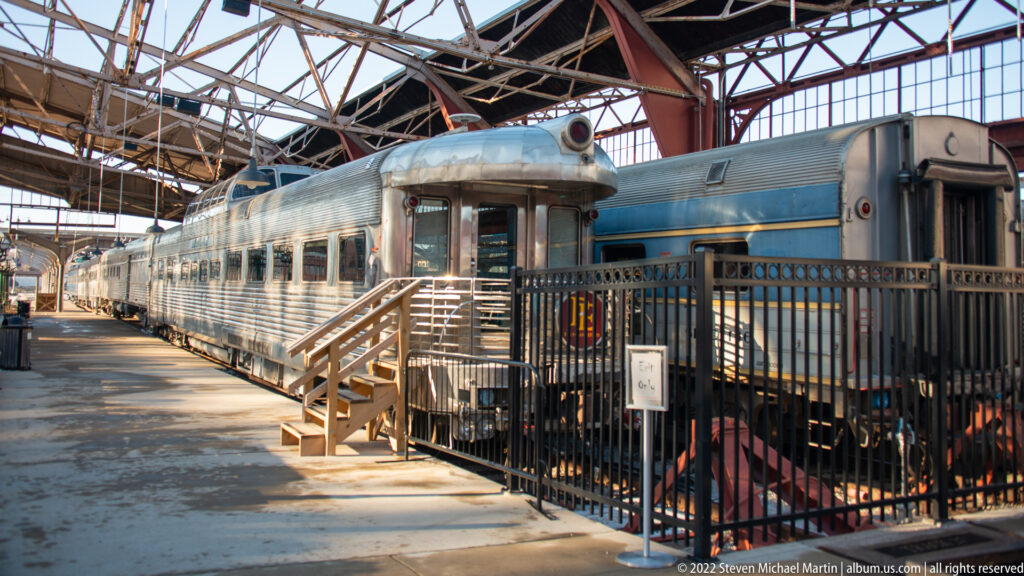 Silver passenger railcars stand behind black railings beneath a large glass roof.