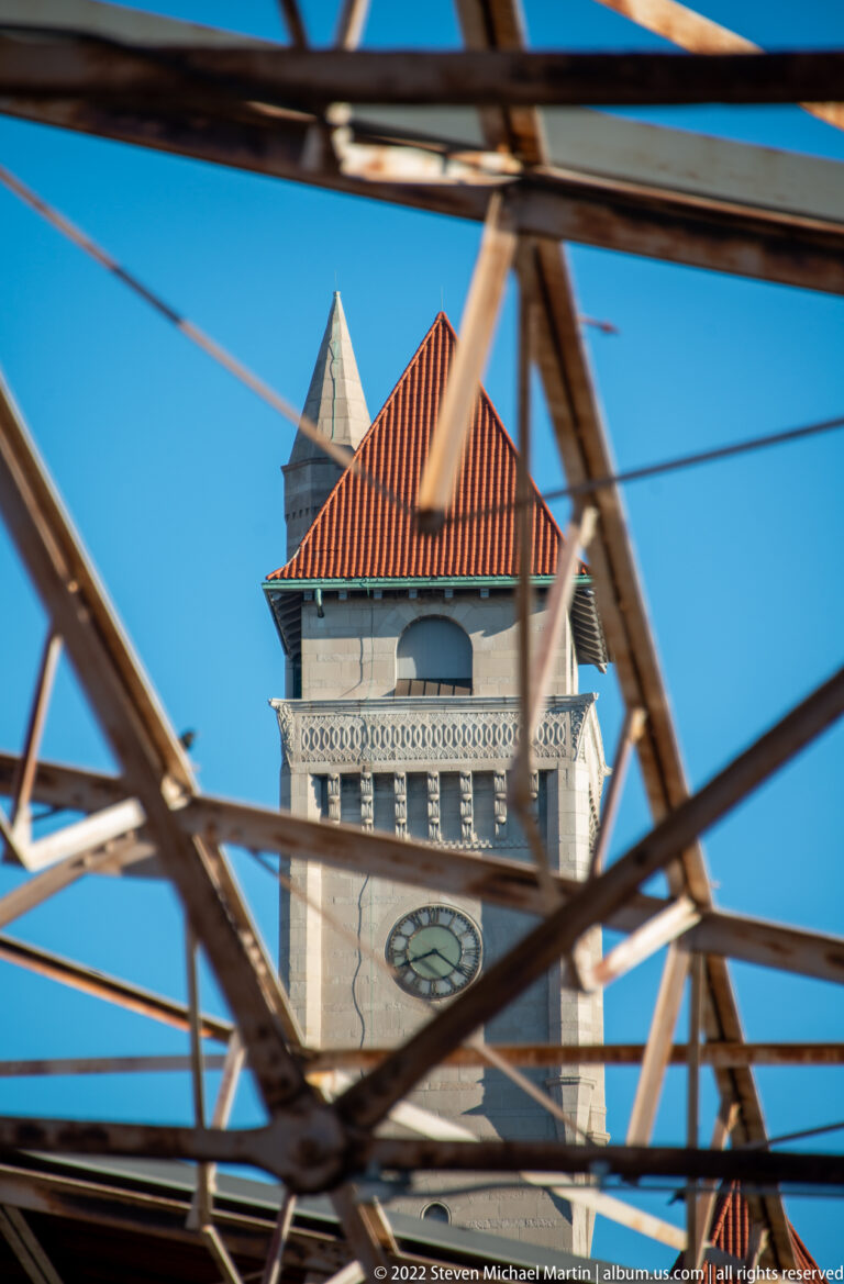 A stone clock tower and red roof appear through intersecting dark steel beams against blue sky.